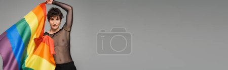 Téléchargez les photos : Brunette bigender personne en haut transparent noir regardant la caméra tout en tenant le drapeau lgbt isolé sur gris, bannière - en image libre de droit
