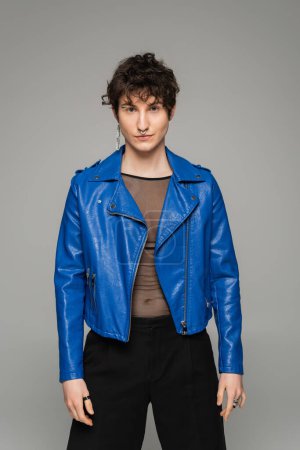 Foto de Modelo pansexual de moda en chaqueta de cuero azul mirando a la cámara aislada en gris - Imagen libre de derechos
