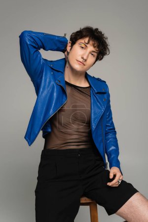 Photo for Fashionable bigender person in blue leather jacket posing with hand behind head isolated on grey - Royalty Free Image