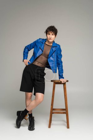 Photo for Full length of bigender person in blue leather jacket and black shorts posing near wooden stool on grey background - Royalty Free Image