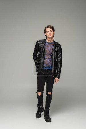 full length of stylish nonbinary person in black leather jacket and ripped pants on grey background