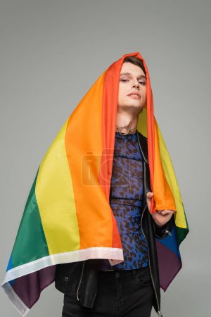 trendige Pangender-Person, die sich mit Regenbogenfahne verhüllt und isoliert auf grau in die Kamera blickt