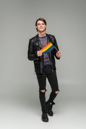 full length of carefree pangender person in black ripped pants and leather jacket standing with small lgbt flag on grey background