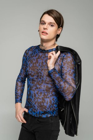 Foto de Persona no binaria de moda con maquillaje que sostiene la chaqueta de cuero negro y mirando a la cámara aislada en gris - Imagen libre de derechos