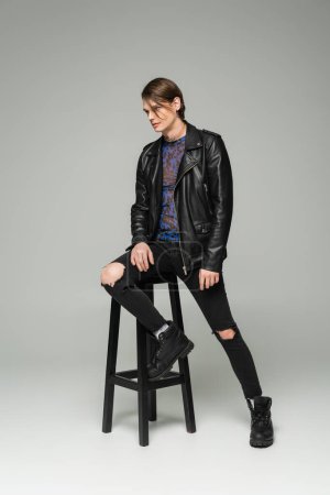 Photo for Full length of bigender model in black leather jacket and ripped pants posing near high stool on grey background - Royalty Free Image