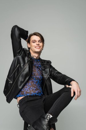 carefree bigender person in black leather jacket sitting with hand behind head isolated on grey