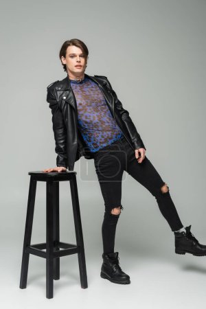 full length of bigender model in leather jacket and ripped pants posing near high stool on grey background
