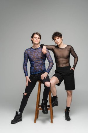 full length of young and fashionable nonbinary models posing near high stool on grey background