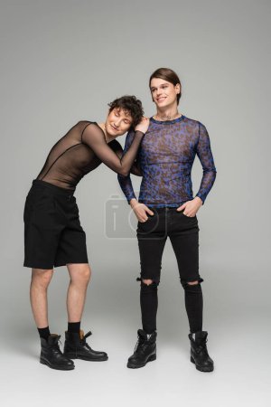 trendige nichtbinäre Person in kurzen Hosen, angelehnt an Partner in Animal Print Top auf grauem Hintergrund