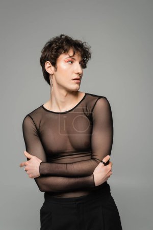 Photo for Young nonbinary person in makeup and black transparent top standing with crossed arms and looking away isolated on grey - Royalty Free Image