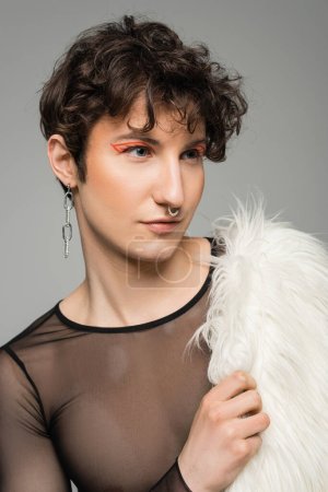 portrait of young pansexual person with makeup holding white faux fur jacket and looking away isolated on grey