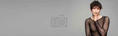 Foto de Alegre modelo pansexual en negro transparente superior mirando cámara aislada en gris, pancarta - Imagen libre de derechos