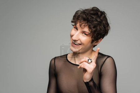 fröhliche bigeschlechtliche Person in schwarzem transparentem Oberteil und silbernen Ringen, die mit dem Finger auf die Kamera zeigen, isoliert auf grau
