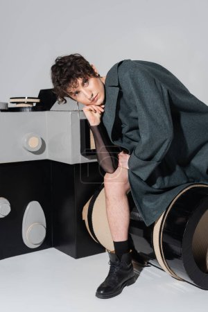 young and dreamy bigender person in coat sitting on model of photo camera on grey background