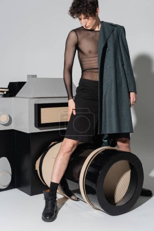 fashionable bigender person in coat and transparent top posing near huge model of photo camera on grey background