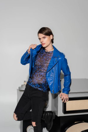 thoughtful bigender person in blue leather jacket looking away near huge model of photo camera on grey background