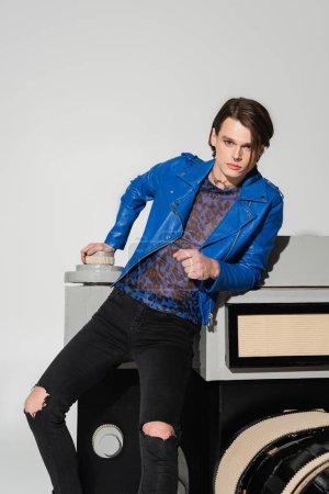 young pansexual person in blue leather jacket and black ripped pants posing near huge model of photo camera isolated on grey