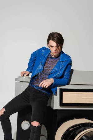 confident nonbinary person in blue leather jacket leaning on model of photo camera isolated on grey