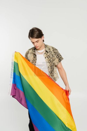 young nonbinary model in animal print blouse holding rainbow flag isolated on grey