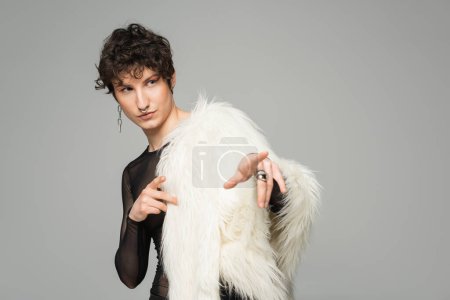 Foto de Modelo no binario de moda en chaqueta de piel sintética blanca apuntando con los dedos y mirando hacia otro lado aislado en gris - Imagen libre de derechos
