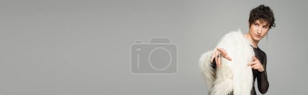 brunette bigender person in white faux fur jacket looking away and pointing with fingers isolated on grey, banner