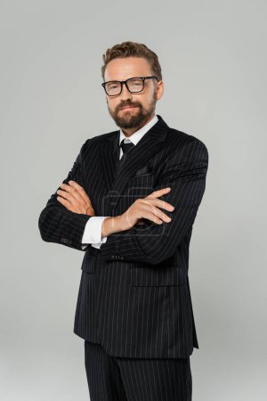 cheerful businessman in formal wear and glasses posing with crossed arms isolated on grey 