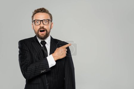 astonished businessman in formal wear and glasses pointing away isolated on grey 