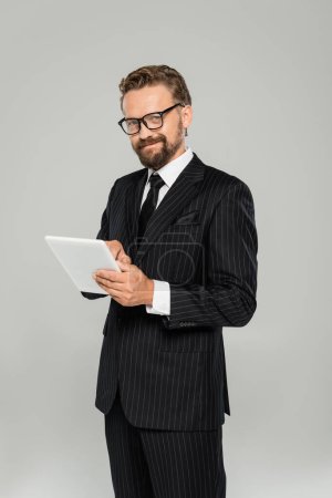 happy businessman in formal wear and glasses using digital tablet isolated on grey 