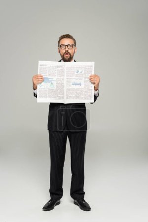 full length of astonished businessman in suit and glasses holding newspaper on grey 