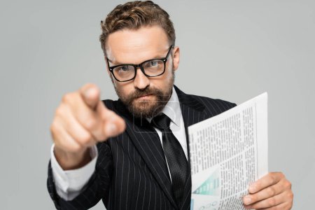 businessman in suit and glasses pointing with finger at camera while holding newspaper isolated on grey 