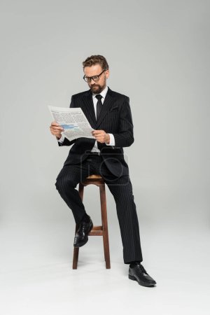 Geschäftsmann in Anzug und Brille liest Zeitung, während er auf einem Hochstuhl isoliert auf grau sitzt 