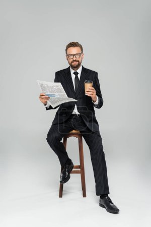 Téléchargez les photos : Pleine longueur d'homme d'affaires heureux en costume et lunettes tenant journal tout en étant assis avec tasse en papier sur gris - en image libre de droit