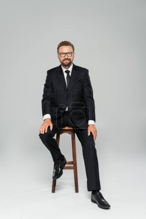 Foto de Longitud completa de hombre de negocios feliz en traje y gafas sentado en la silla alta de madera en gris - Imagen libre de derechos