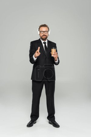 longitud completa de empresario feliz en gafas y traje sosteniendo taza de papel y señalando con el dedo en gris 