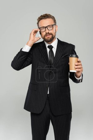Geschäftsmann in Brille und Anzug spricht auf Smartphone und hält Pappbecher isoliert auf grau 