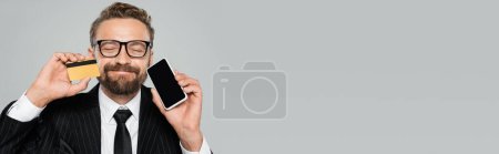 joyful businessman in suit and glasses holding smartphone and credit card isolated on grey, banner 