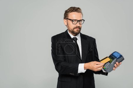 successful businessman in suit and glasses holding credit card near payment terminal isolated on grey