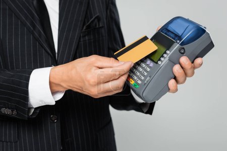 Téléchargez les photos : Vue recadrée d'un homme d'affaires en costume tenant une carte de crédit près d'un terminal de paiement isolé sur gris - en image libre de droit