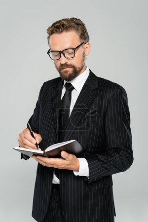 gut gekleideter Geschäftsmann mit Brille und formaler Abnutzung Schrift in Notizbuch isoliert auf grau 