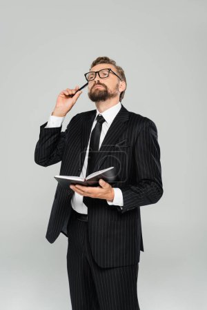 Nachdenklicher Geschäftsmann mit Brille und formeller Kleidung, Notizbuch isoliert auf grau 