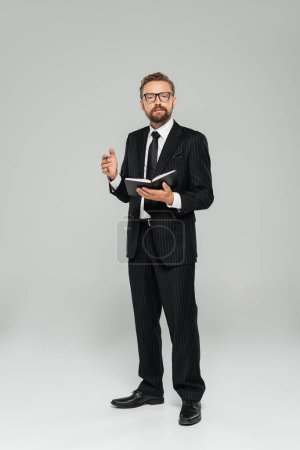 full length of well dressed businessman in glasses and formal wear holding notebook on grey 