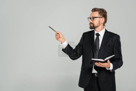 well dressed businessman in glasses and formal wear holding notebook and pointing with pen isolated on grey 