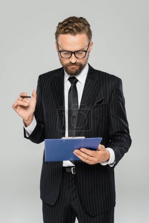 well dressed businessman in glasses and formal wear holding clipboard with pen isolated on grey 
