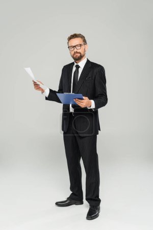 full length of well dressed businessman in glasses holding clipboard and paper on grey 