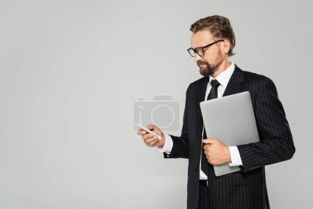 homme d'affaires bien habillé dans des lunettes tenant ordinateur portable et utilisant smartphone isolé sur gris 