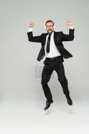 Foto de Longitud completa de empresario excitado en gafas y auriculares saltando en gris - Imagen libre de derechos