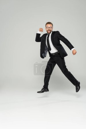Aufgeregter Geschäftsmann in Brille und Anzug springt auf grau 