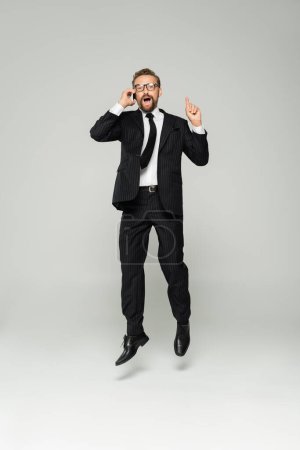 full length of excited businessman in glasses talking on smartphone and pointing up while jumping on grey 