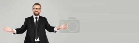 happy businessman in suit and glasses looking at camera and showing welcoming gesture isolated on grey, banner 