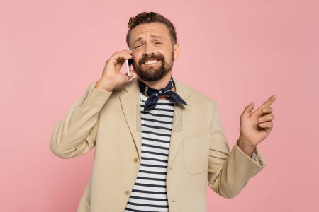 cheerful french man in blazer and neck scarf talking on smartphone while pointing away isolated on pink 